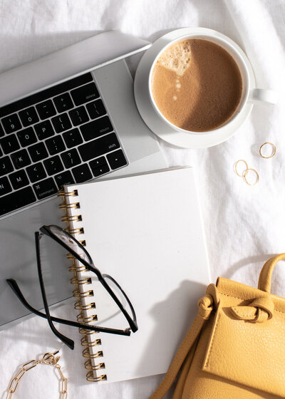 Laptop, coffee, notebook, glasses, and gold rings on a white surface.