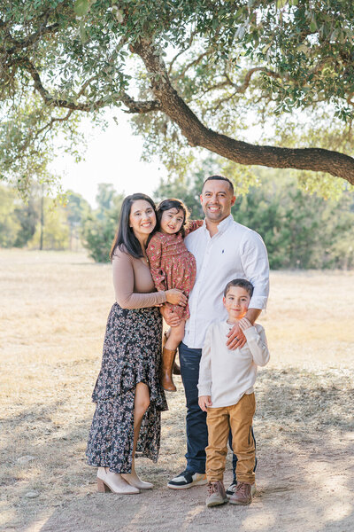 A family of four stand under a tree at Brushy Creek Lake Park. The father holds the daughter, the son stands to the side of his father, and the mother wraps her arms around her daughter. They all look at the camera smiling.