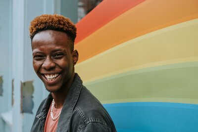 This image shows a masculine-presenting person of color, smiling over their left shoulder toward the camera. They are standing against a rainbow-painted mural.