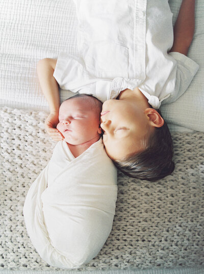 Big brother lies eyes-closed on bed next to his sleeping newborn brother