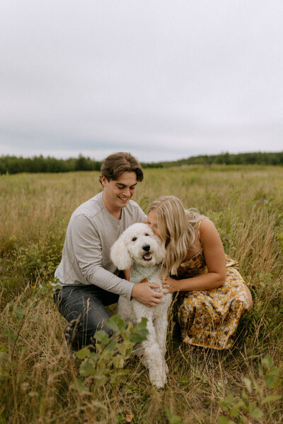 Engaged couple kneeling down kissing dog in field