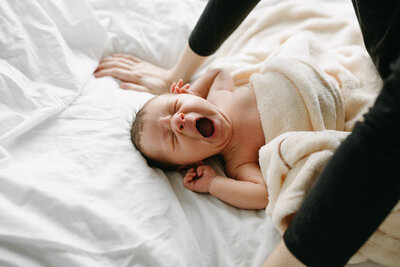 Newborn yawning and stretch with soft natural light spilling in on the bed. A mother gently hovering over her baby 