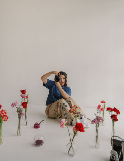 A photographer sits on the floor with a camera, surrounded by various flowers in glass vases. The setting is bright and minimalistic, conveying creativity.