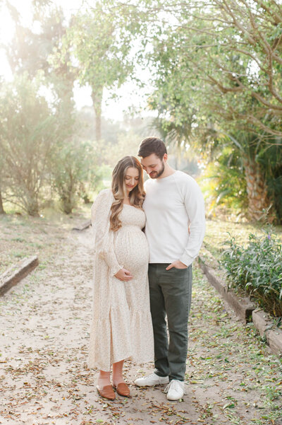 Soon to be parents stand in a beach access pathway, mom holding her belly and both parents staring at moms belly thinking of their new chapter together.