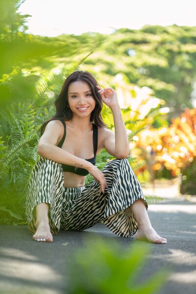 Girl with dark hair in black shirt and zebra pants sitting on ground smiling at camera, surrounded by greenery.
