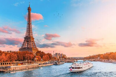 The iconic Eiffel Tower stands tall against a pastel-hued sunset sky, framed by autumn-colored trees along the Seine River. A sightseeing boat glides through the sparkling water, capturing the romance and charm of Paris as a dream destination supported by vacation fundraising for charities.