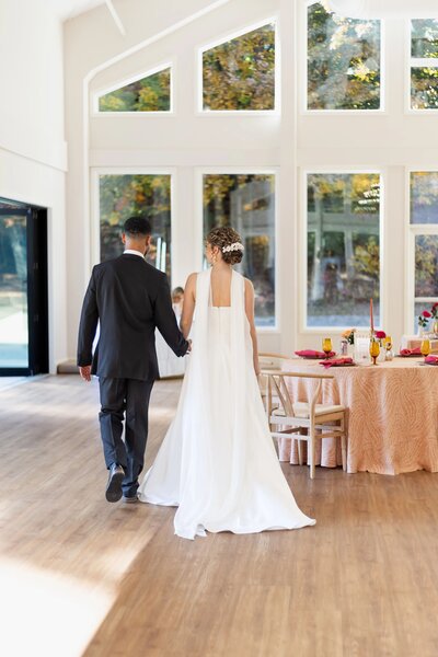 Best Rhode Island Wedding Photographers | Kelsey Sheehan Photography Timeless Rhode Island Weddings | A bride and groom walk hand in hand toward a sunlit table in an elegant room with large windows, conveying a serene and joyful wedding atmosphere.