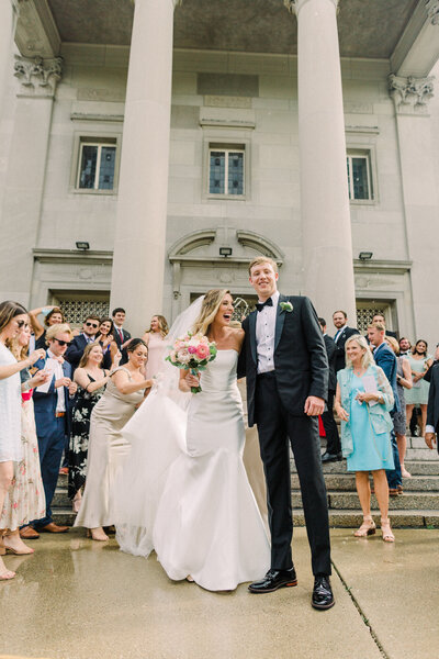 A candid wedding photo of a bride and groom walking outside the church as guests cheer and celebrate, lined up on the steps in a beautiful post-ceremony moment.