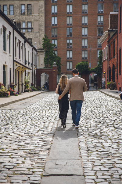 Engaged couple walking hand in hand down cobblestone street in historic Philadelphia neighborhood