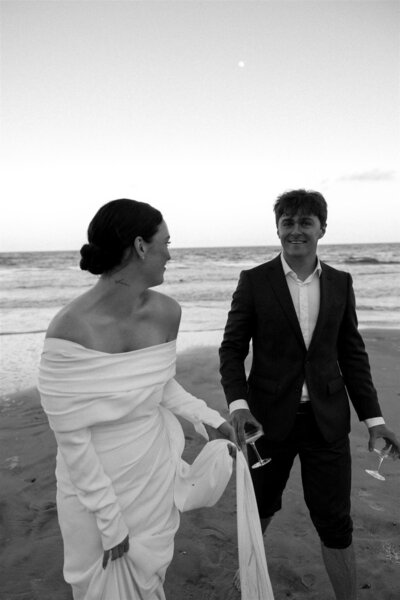 A bride and groom laugh together while walking up the beach holding martinis