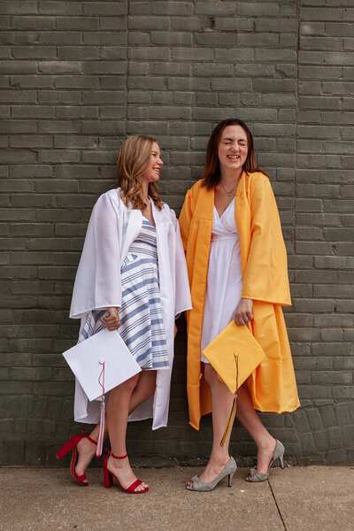 Image of 2 grads in different caps and gowns, laughing together while leaning against a wall