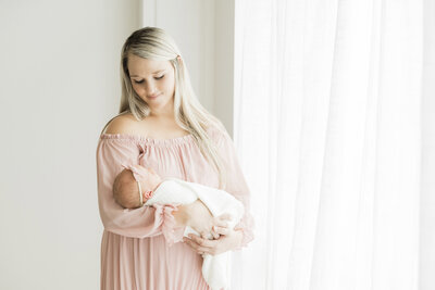 Mother holding her swaddled newborn in a soft pink dress during a natural light studio session with a Kansas City newborn photographer.