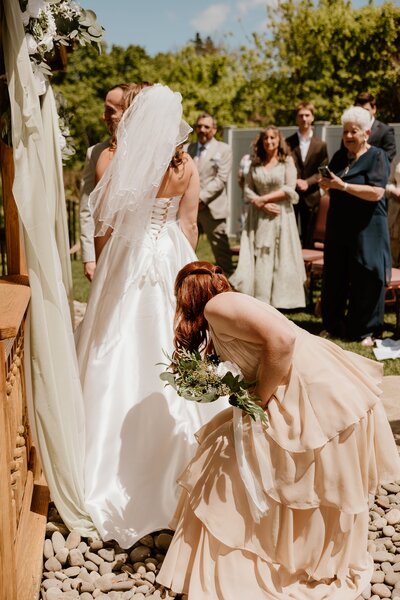 Bridesmaid adjusts the bride’s train during an intimate Westchester outdoor wedding ceremony, surrounded by guests on a sunny spring day.