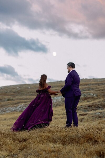 Bride and groom hold hands as moon rises behind them.