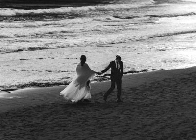 a black and white image at the end of their sunset elopement package, a bride and groom in their wedding attire hold hands as they stroll along the shoreline of a beach