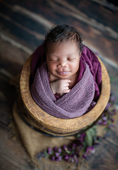 Black haired smiling baby girl wrapped in deep purple sitting in rustic bucket surrounded by purple flowers 