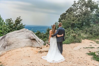 A bride and groom stand looking out at a mountain overlook after eloping. Their arms are wrapped around each other and their backs face the camera. They are standing on a dirt clearing with a large rock nearby and lush green trees. The bride is wearing a fitted white wedding gown and the groom is wearing a navy blue suit.