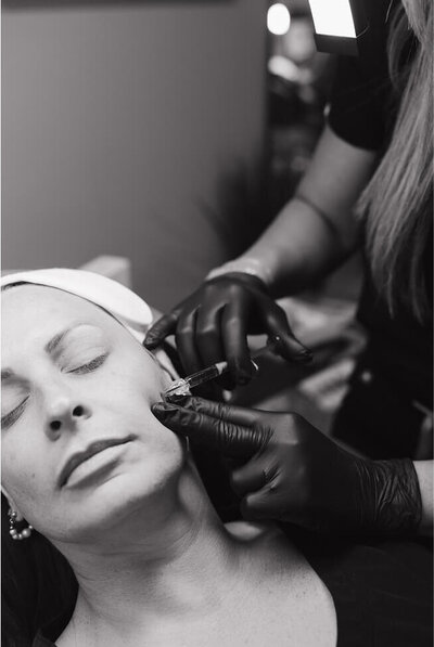 A woman is getting aesthetic injectables in her cheeks. The nurse is gently injecting the Botox into her cheek.