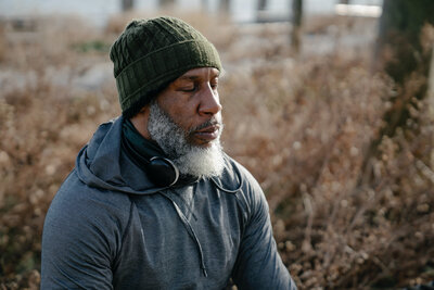 A masculine presenting person of color, with a gray beard and a green beanie, sits on the ground. He faces past the camera with his eyes closed, headphones around his neck.