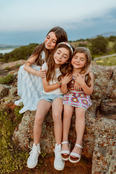 Three sisters hug each other on a rock with their eyes closed with Kansas City Photographer.