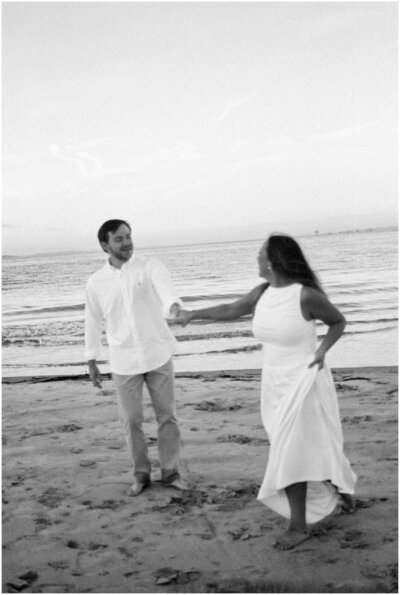 A woman in a white dress leads her fiancee down the beach by the hand as they both laugh