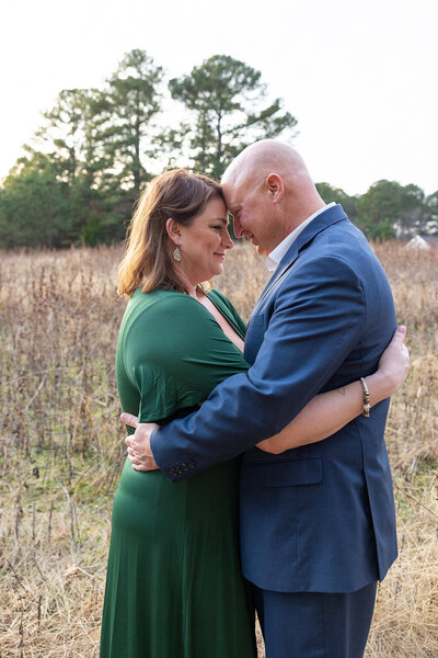 Bride to be with green flowing dress hugs groom in blue suit in a field