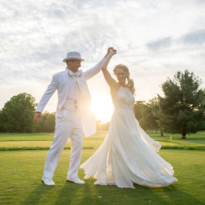 groom in white twirls bride in front of the setting sun