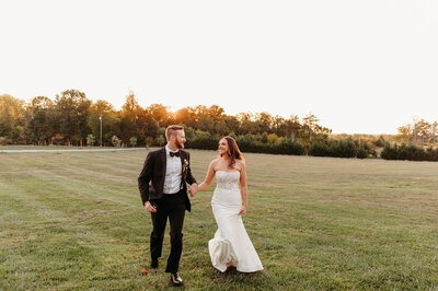 A couple, dressed in wedding attire, walks hand in hand across a green field during sunset at a picturesque Leesburg VA wedding venue. The groom wears a black suit and bow tie, while the bride is in a white gown. The setting sun casts a warm glow over the romantic scene.