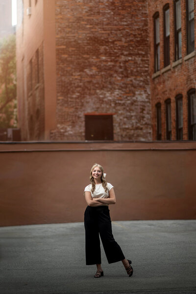 A high school senior in black pants and white top stands with arms crossed in a back alley