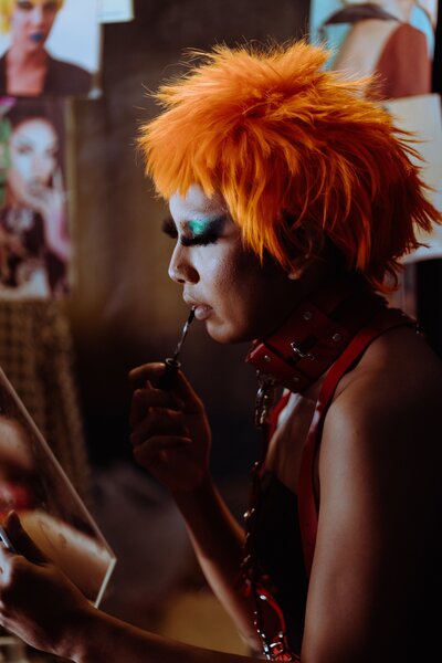 A person with bright orange hair and glittery green eyeshadow looks past the camera into a mirror. They are wearing a leather choker and chain, while applying lipgloss.