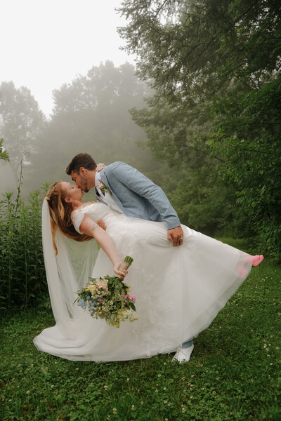 Bride and groom sharing a romantic kiss, with the bride holding a bouquet in a misty, scenic outdoor setting.