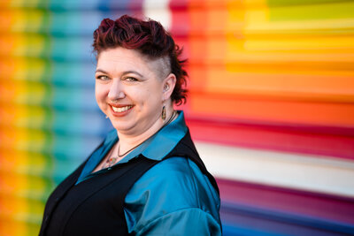 A person smiling while standing in front of a rainbow colored wall.