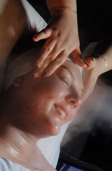 Brittney is giving her client a facial with her hands and smiling. Her client is also smiling while getting the facial and looks pleased