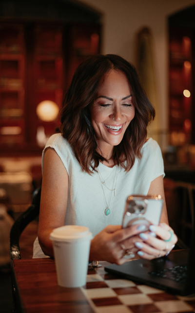 brunette woman smiling looking at phone in a dark woody background and a coffee cup next to her