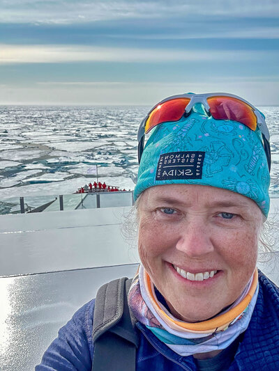 Person smiling on a ship deck wearing a blue headband, sunglasses, and a scarf, with icy sea and distant people on the bow in the background.
