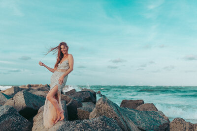 Woman in a glittering gown poses confidently on rocks by the ocean for a glamorous beach portrait in St. Augustine.