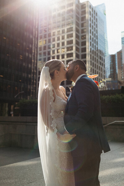 A bride and groom share a kiss outdoors in a city setting, surrounded by tall buildings and sunlight streaming from above. Captured beautifully by an Oshawa wedding photographer, the moment is both romantic and timeless.