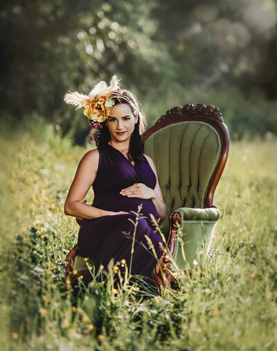 Pregnant woman in purple dress embraces her belly while sitting in green velvet victorian chair in field