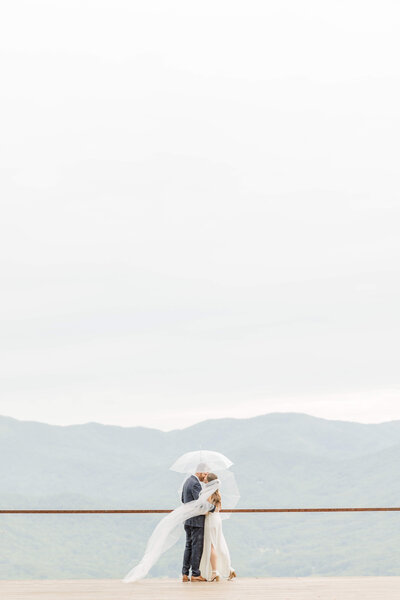 Bride and groom share a kiss under umbrellas with mountains in the background during their rainy day elopement, captured by Destination Wedding Photographer.