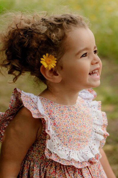 Close up side profile image of a young girl stood a field grinning with a flower in her hair