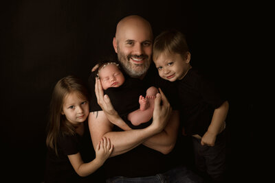 A proud father holds his newborn baby while siblings lovingly pose beside him during a dark-toned, elegant studio session by Duehr Photography in Maple Grove, Minnesota