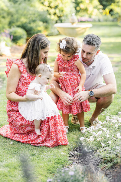 A mother holding a young toddler girl in a sun light rose garden as the toddler admires a soft pink rose she holds in her hand. 