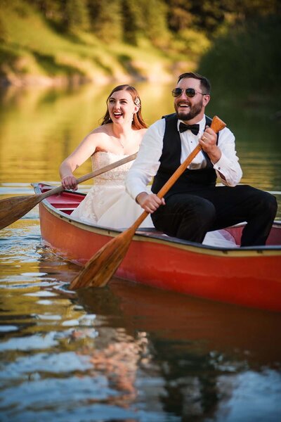 A bride and groom laugh as they paddle a canoe through a river in Whitefish, Montana.