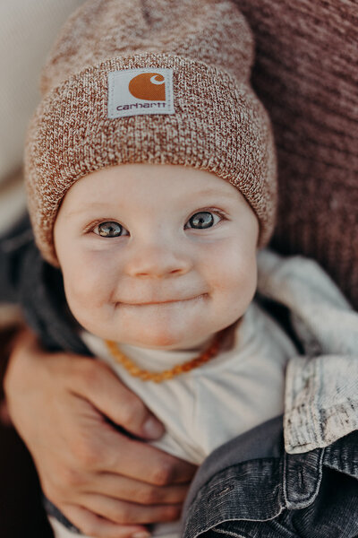 A six month old adorable baby boy sucks his lips in while he smiles, with bright blue eyes wearing fall clothing, including a hooded denim jacket and a Carhartt beanie.