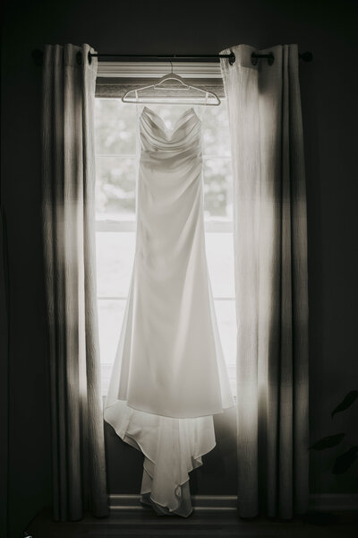 A simple, elegant white wedding dress hangs from a hanger in front of a softly lit window framed by sheer curtains.