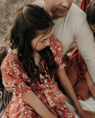 A teenage girl lays back on her dad while wearing a beautiful red floral dress as he prays over her sitting on a blanket outside in Denver Colorado.