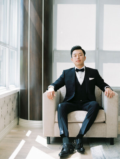 handsome asian groom in tuxedo sitting in a linen club chair