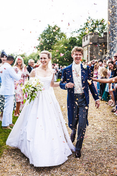 Talulah and Thomas walk hand in hand, smiling at guests as they are showered with confetti at a church wedding