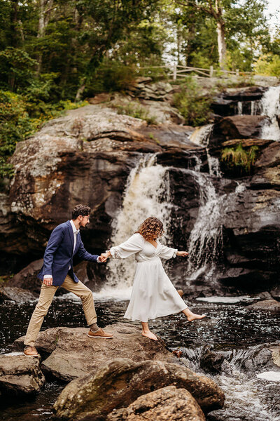 Bride and groom walk hand in hand across rocks in front of waterfall