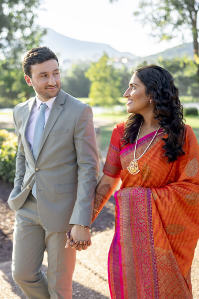 A couple holds hands while walking outdoors. The man wears a light gray suit and tie, while the woman wears a vibrant orange sari with pink details. They are surrounded by greenery, with a sunny sky and distant mountains in the background.